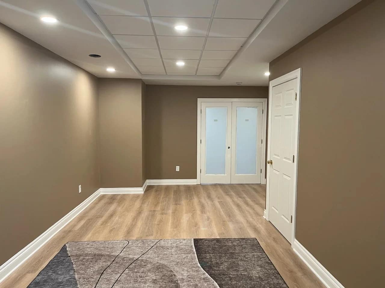 A basement room with brown walls, light wood flooring, recessed ceiling lights, and a section of a gray and beige area rug. At the far end are two frosted glass double doors and a white door on the right.