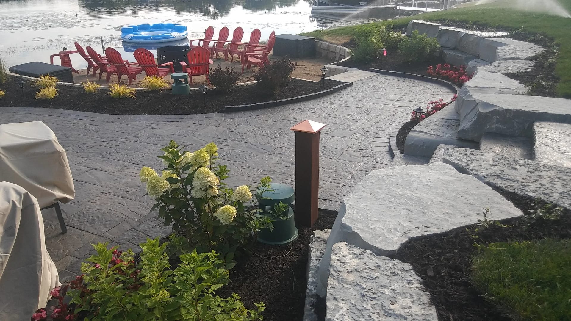 A landscaped lakeside patio with large stone slabs, flower beds, a paved walkway, red Adirondack chairs around a fire pit, and a blue plastic pool by the water.